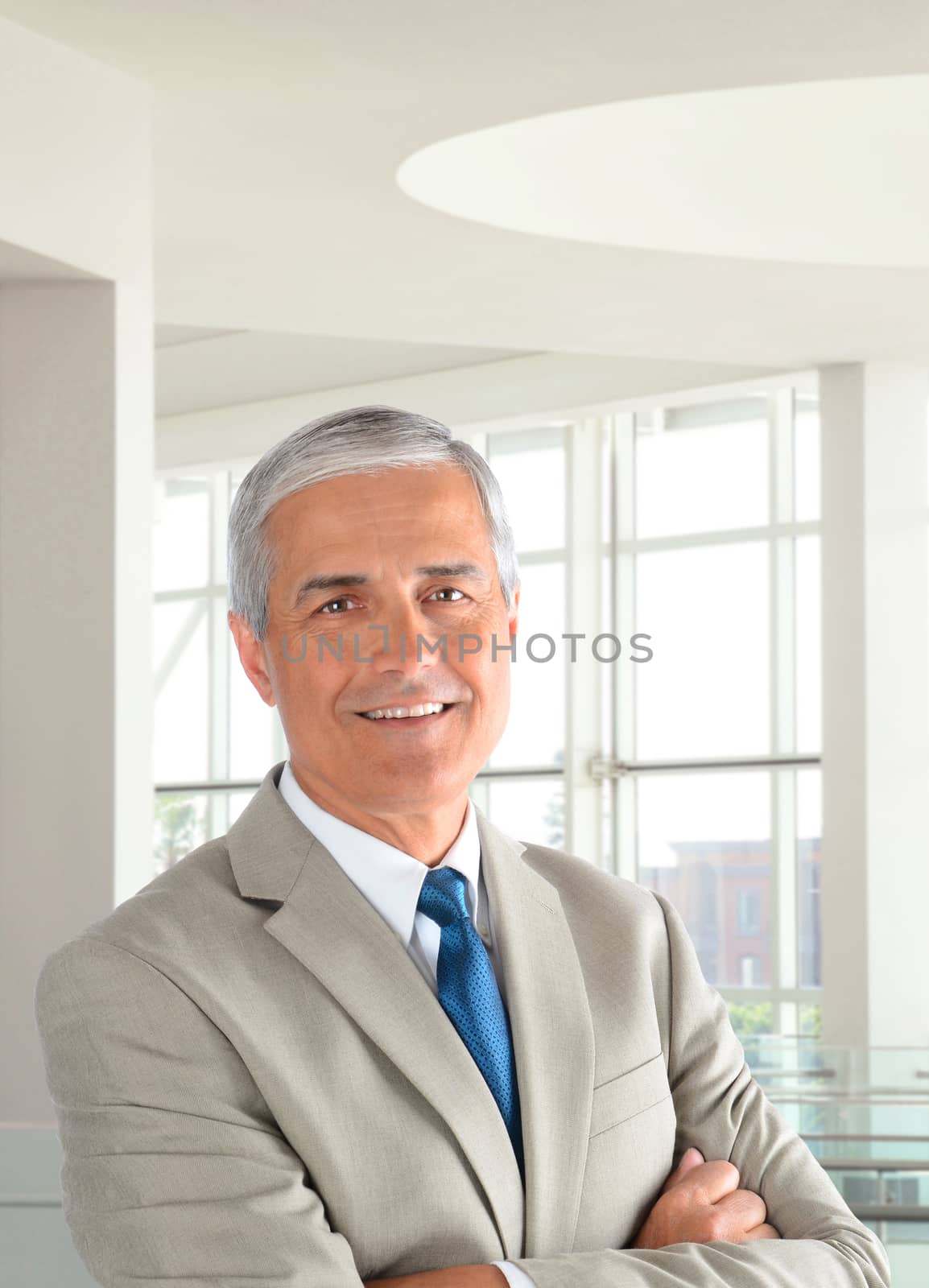 Portrait of a middle aged businessman wearing a light tan suit with his arms folded in a modern office setting. Vertical format, with the man smiling.