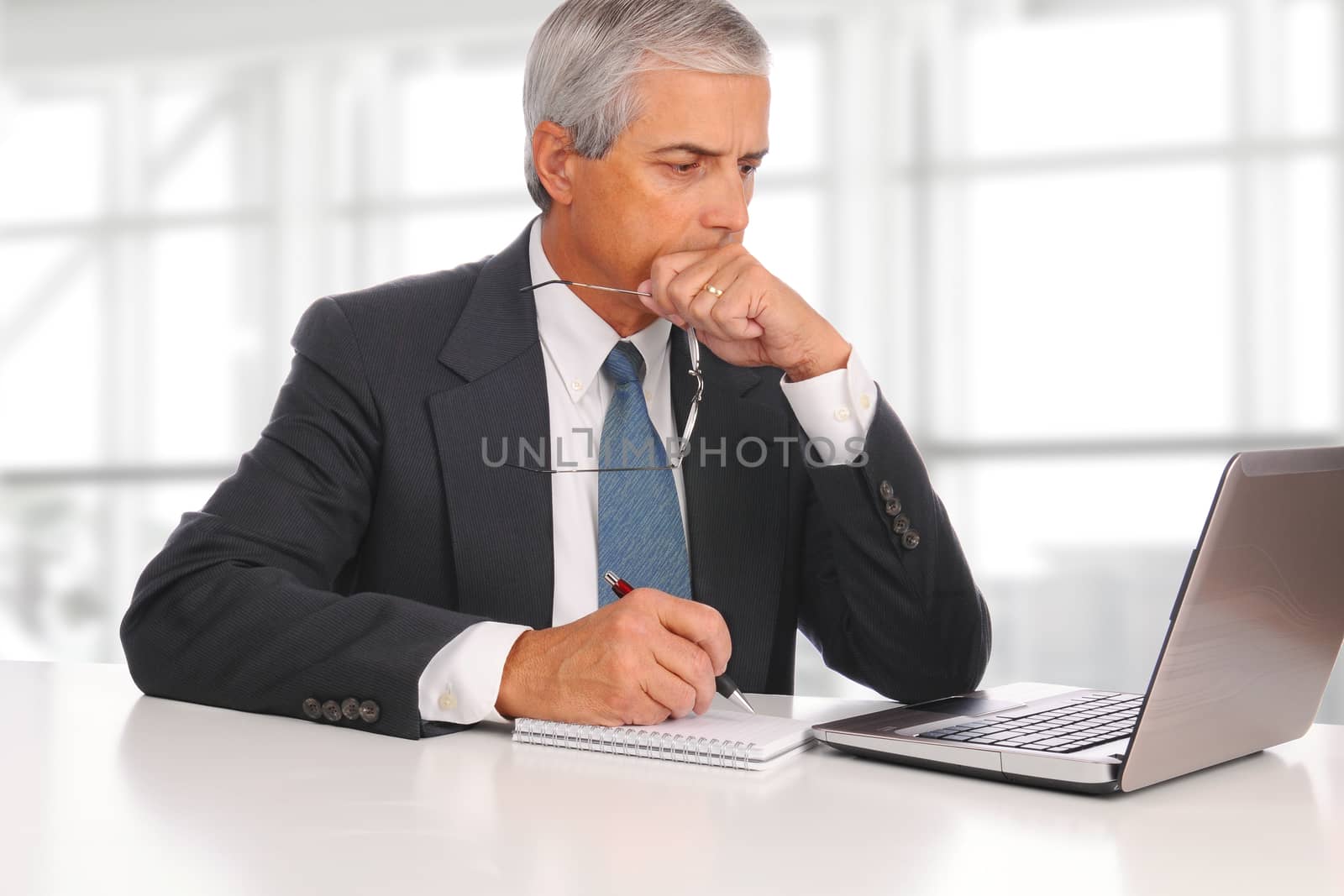 Mature businessman seated at his desk in a modern office building. In front of him is a laptop and note pad. The man is deep in thought.