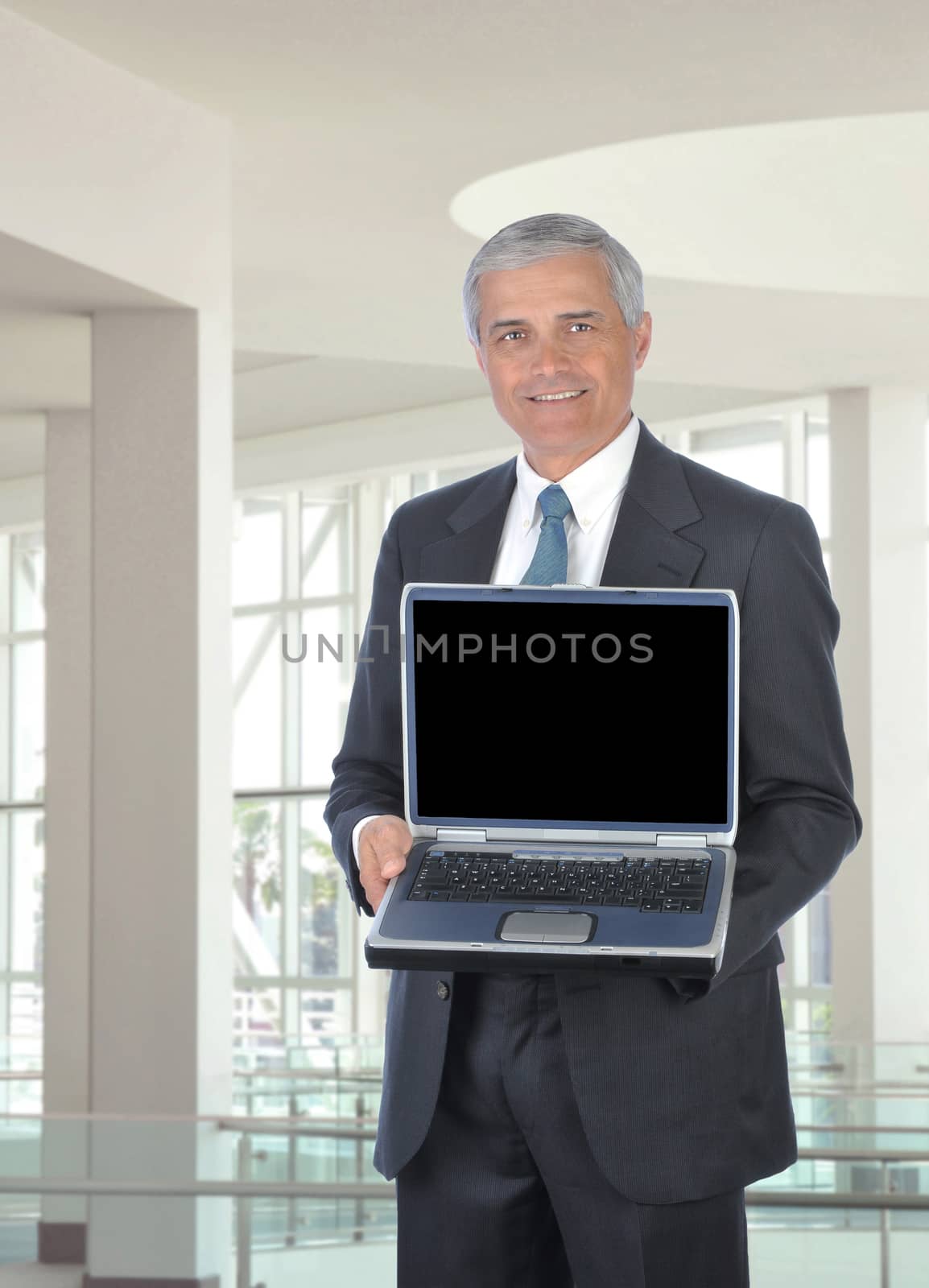 Smiling middle aged businessman holding an open laptop computer. Man is standing in the atrium of a modern office building. Vertical format.