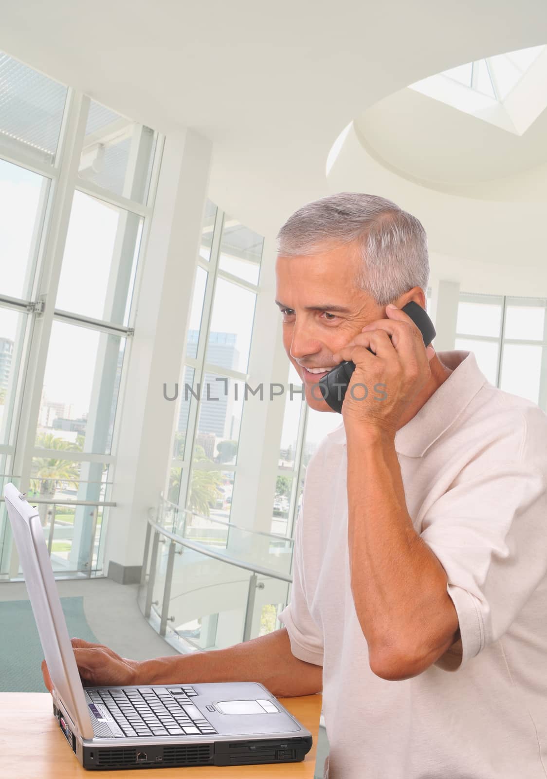 Middle aged Businessman in modern office setting talking on his cell phone and seated at his laptop computer.