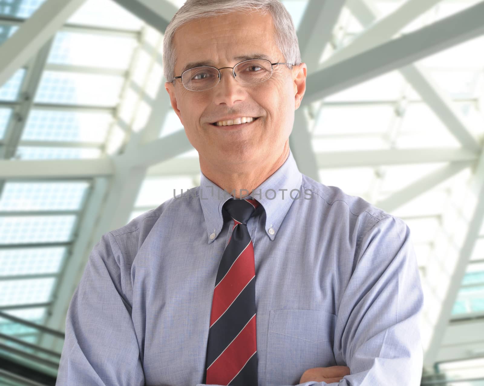Middle aged businessman standing in modern office lobby with his arms crossed. Close up in square format with man smiling at the camera.