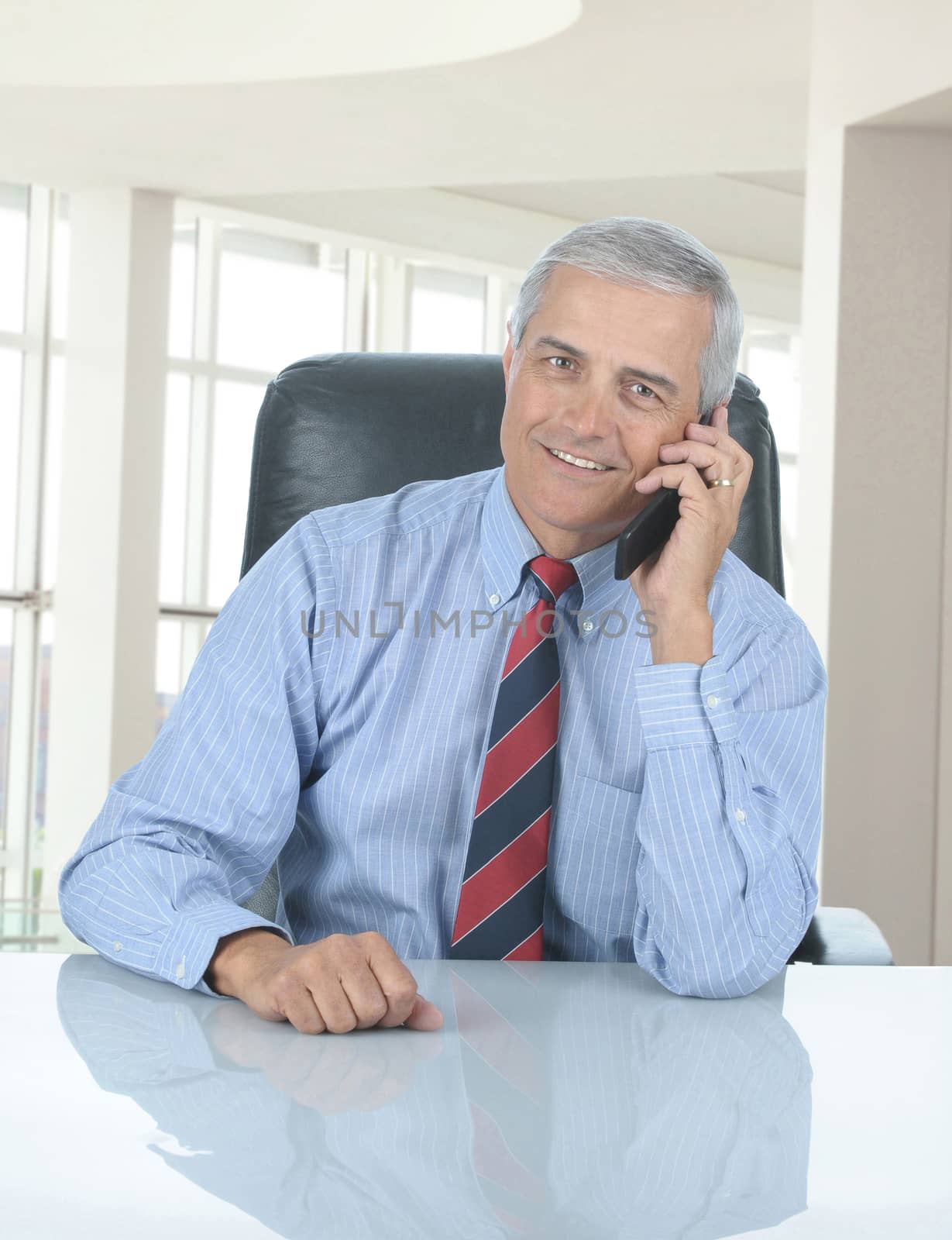 Middle aged businessman seated at his desk in modern office talking on his cell phone. Vertical format with man reflection in desk.