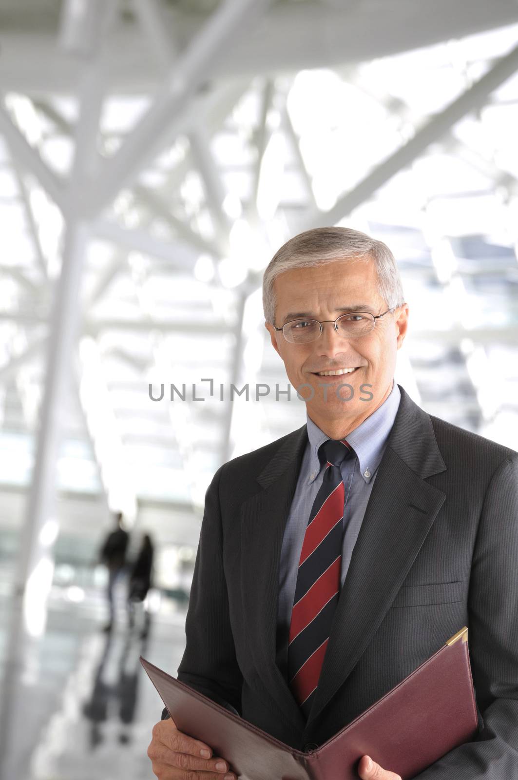 Closeup of a mature businessman inside a modern office building. The man is holding a leather portfolio and smiling at the camera. Blurred people are in the background.