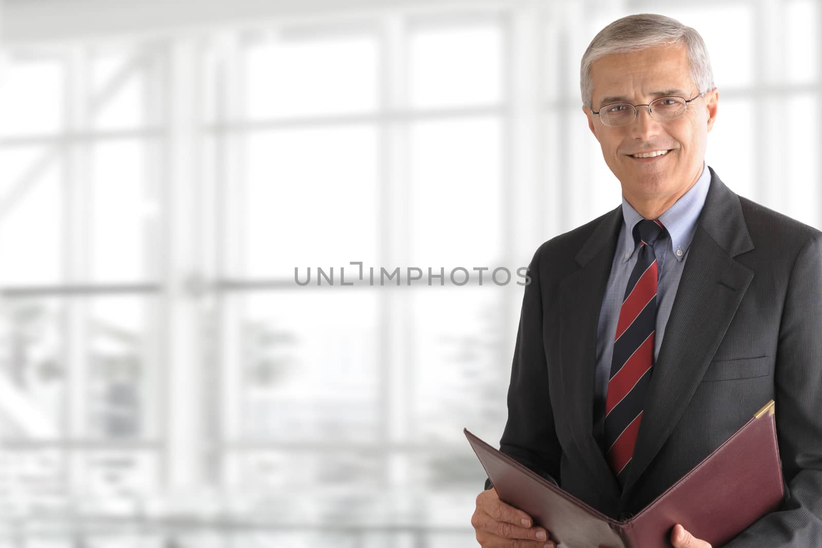 Mature smiling business manager holding a folder while standing in front of a large office window. Horizontal format with copy space.