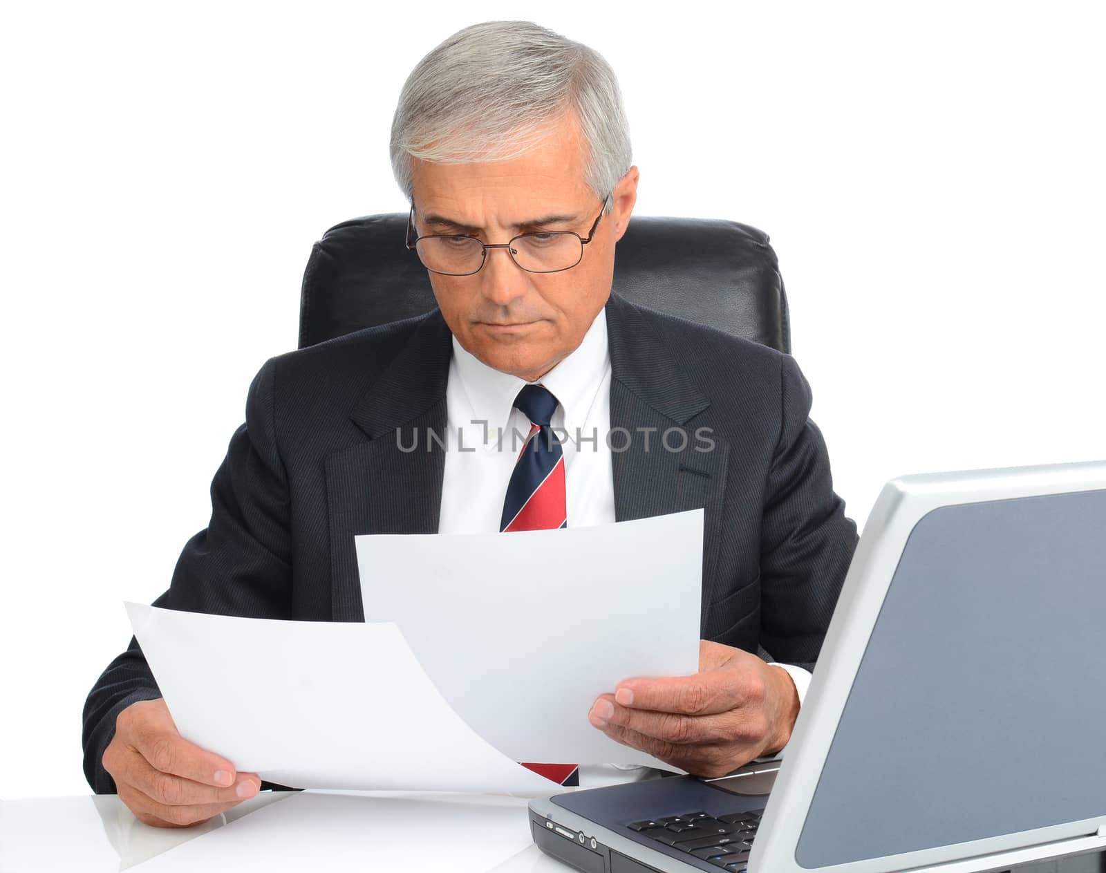 Mature businessman at desk reading papers. Man is wearing eye glasses and has a laptop computer.