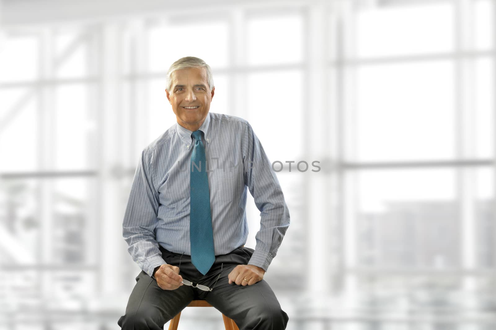 Portrait of smiling senior businessman sitting on a stool against office window background while looking at camera.