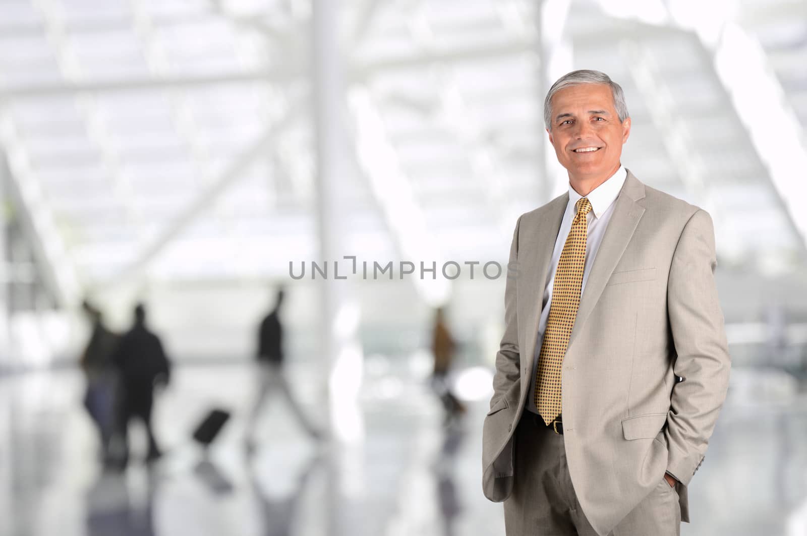Mature businessman in airport concourse with blurred travellers in background. Man has hands in his pockets and is smiling. Horizontal Format