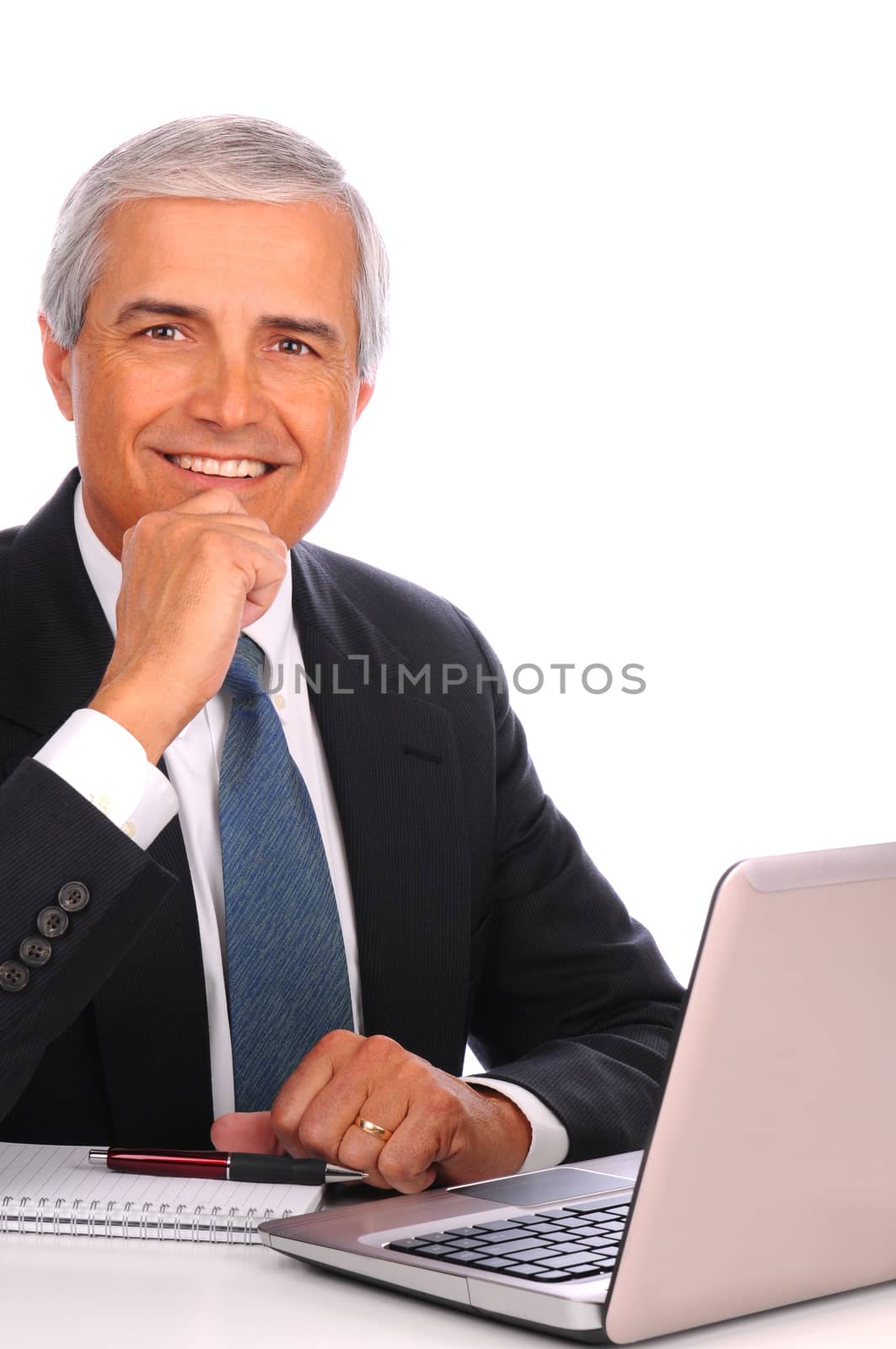 Middle aged businessman seated at his desk in front of laptop computer. Man is smiling with one hand at his chin. Vertical format over white a background.