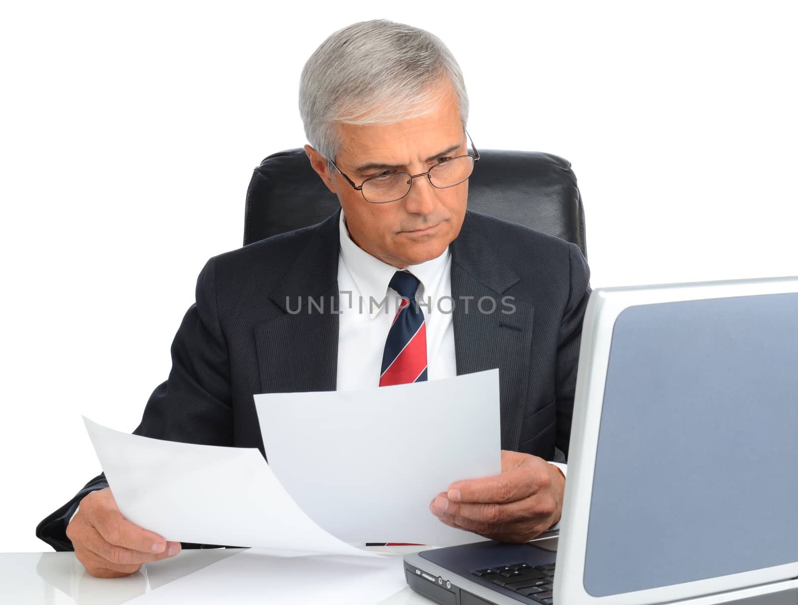 Mature businessman at desk comparing note to laptop screen. Man is wearing eye glasses over a white background.