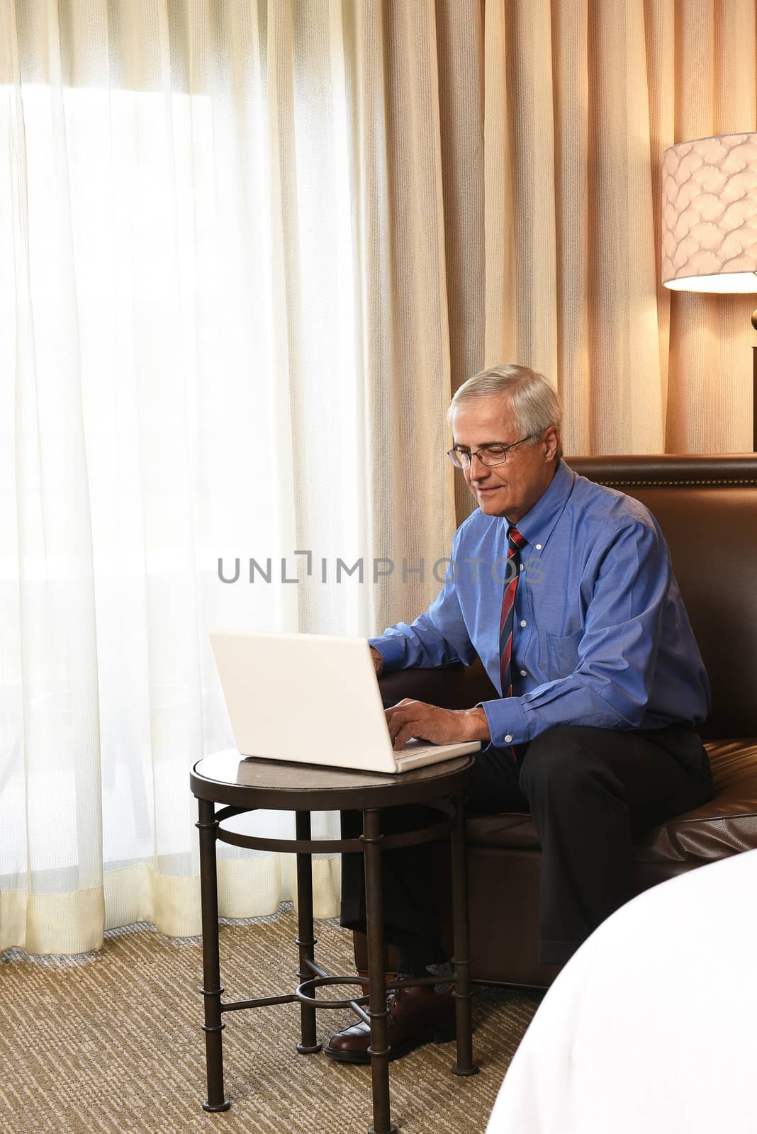 A senior businessman seated on the couch of his hotel room working on his laptop computer on a small table in front of him.