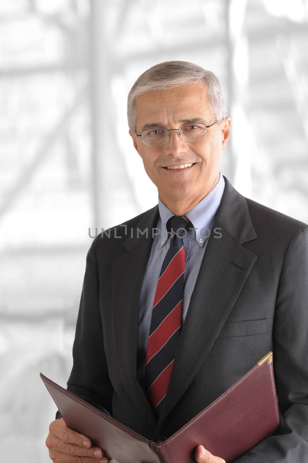 A mature businessman with a leather folder in a modern office building. The background is out of focus and high key.