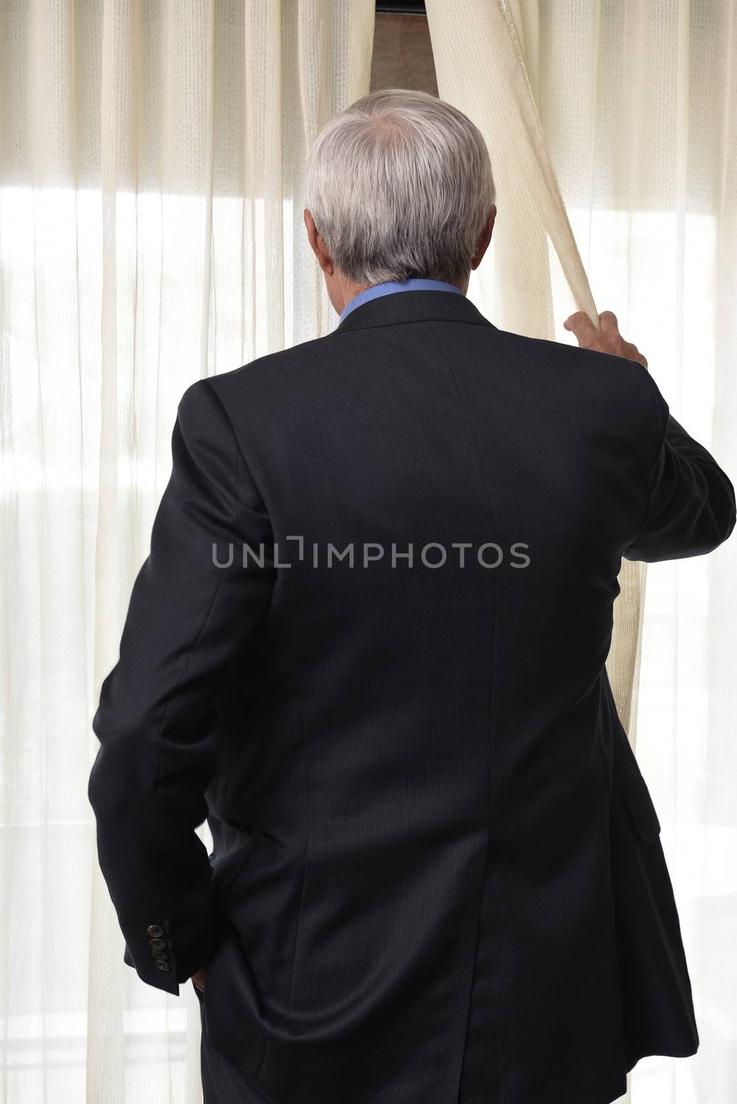 Closeup of a man seen from behind pulling back the drapes and peeking out a window.