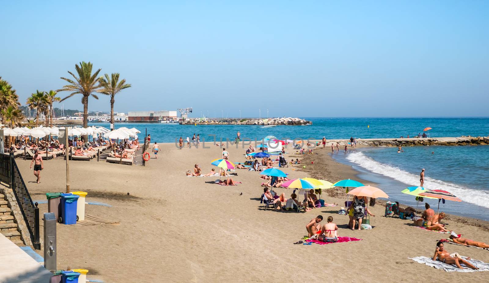 Marbella, Spain - June 27, 2018. People at the beach with palm trees on the shore of the Mediterranean Sea, Marbella city and resort area, province of Malaga, Andalucia region, Spain, Western Europe.