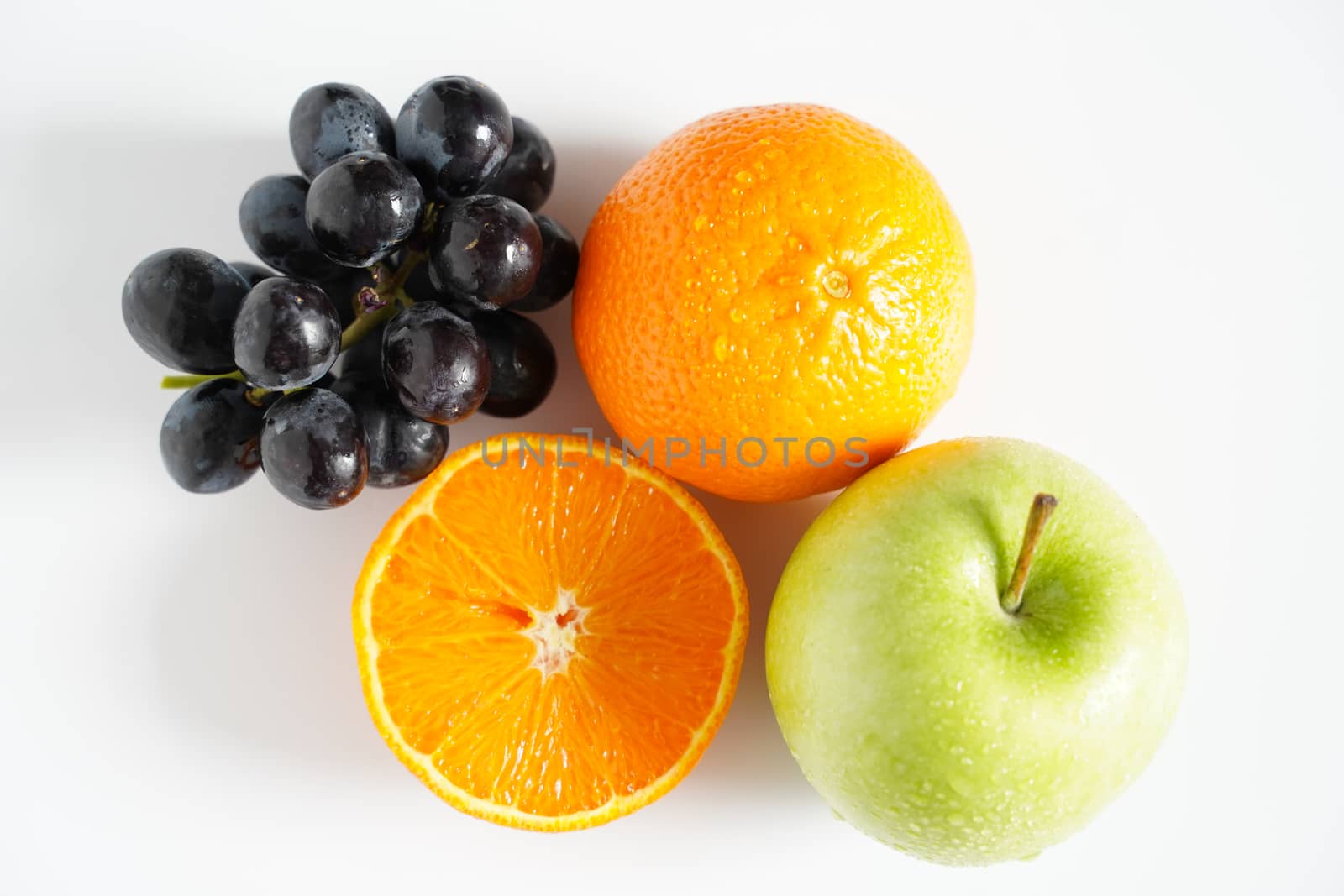 A granny smith green apple, some black grapes and an orange against a plain whit background
