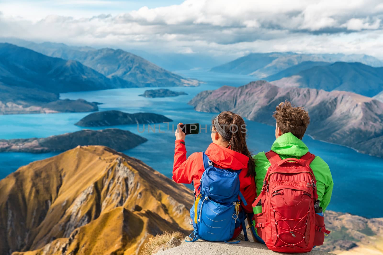 Hikers looking at view on mountain top summit on hiking travel vacation - couple taking photo with phone. Wanderlust adventure people relaxing. Summit of famous hike to Roys Peak, New Zealand by Maridav