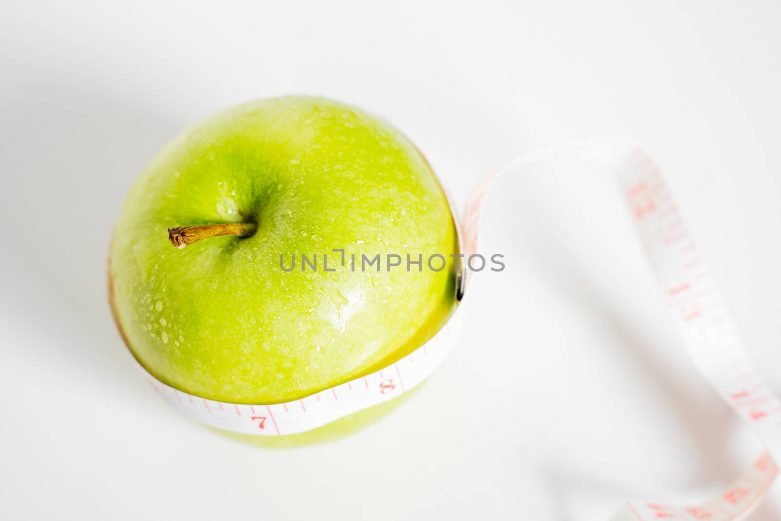 A granny smith green apple with a measuring tape wrapped around it against a plain white background