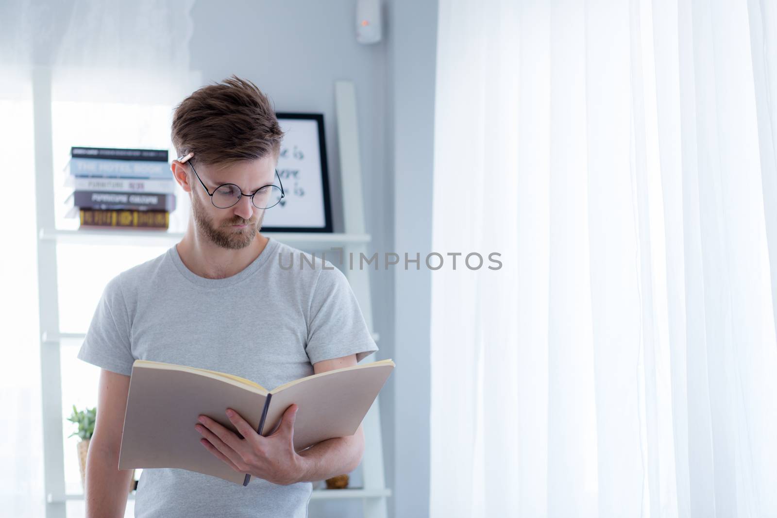 Handsome guy in eyeglasses is reading book preparing exam with standing at the living room - education concept.