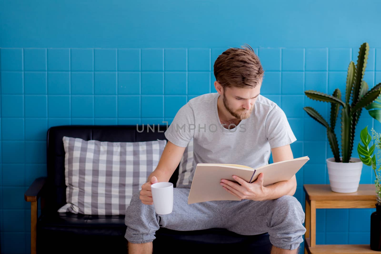 Young man reading book and drinking coffee on sofa in the living room, education concept