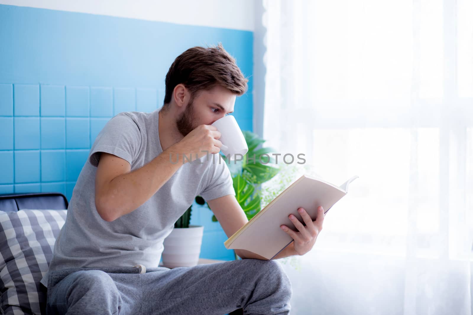 Young man reading book and drinking coffee on sofa in the living room.