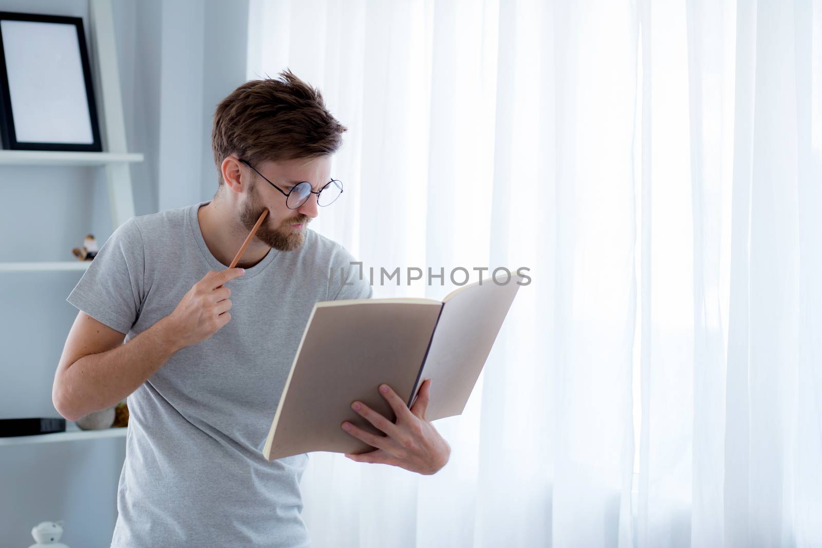 Handsome guy in eyeglasses is reading book preparing exam with standing at the living room, education concept