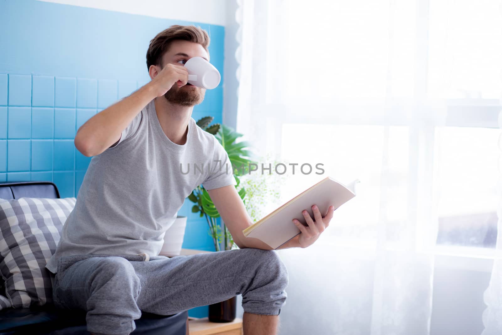 Young man reading book and drinking coffee on sofa in the living room.