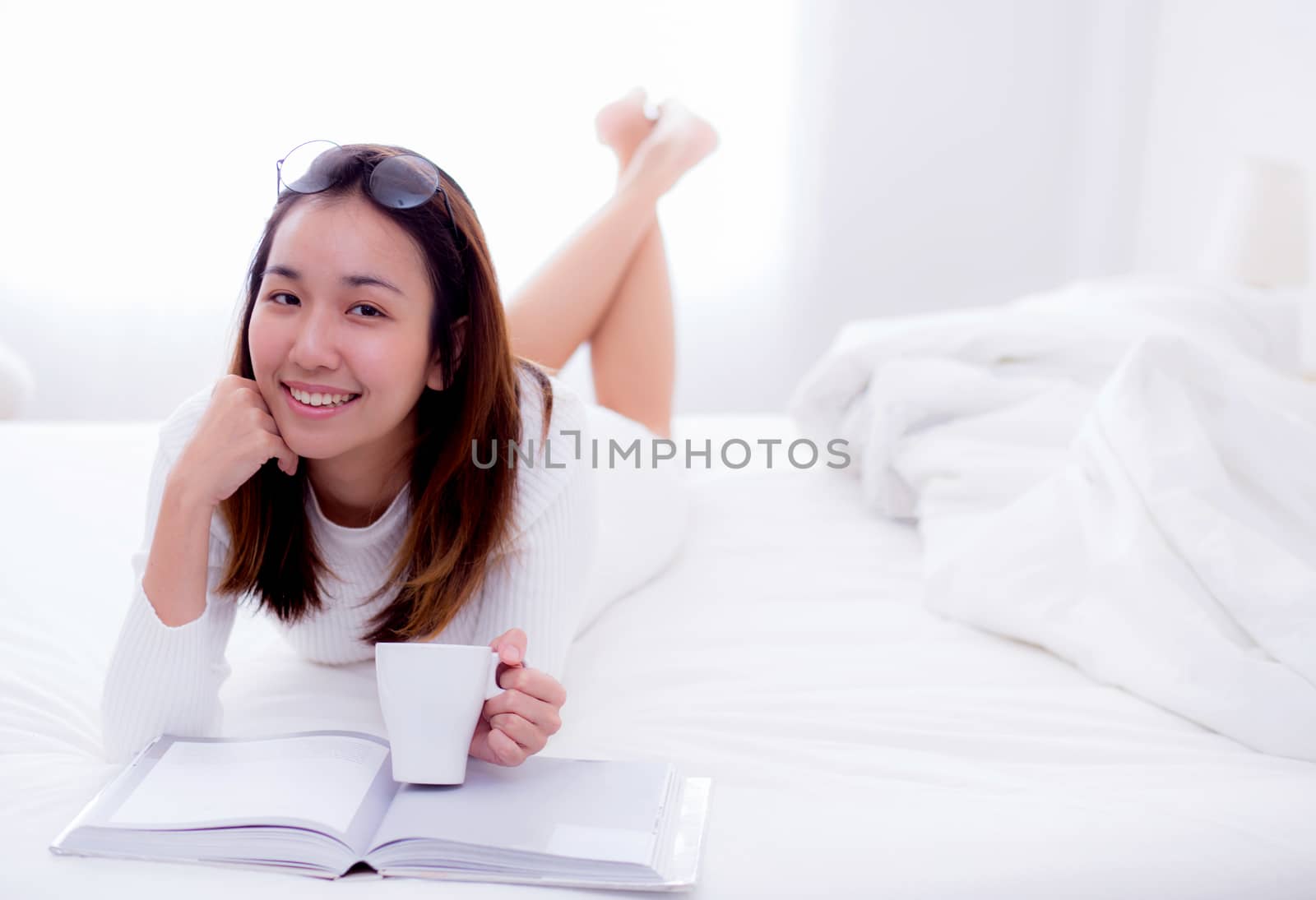 close up of a charming woman lying on bed reading a book and holding coffee cup looking into camera in bedroom.