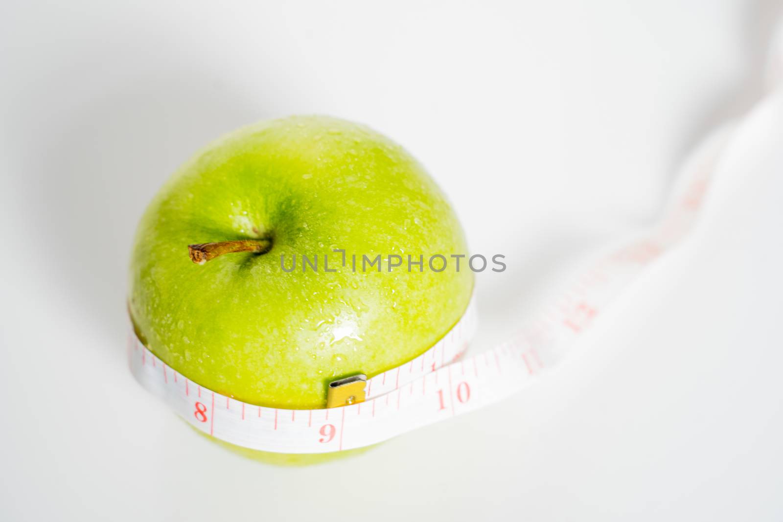 A granny smith green apple with a measuring tape wrapped around it against a plain white background