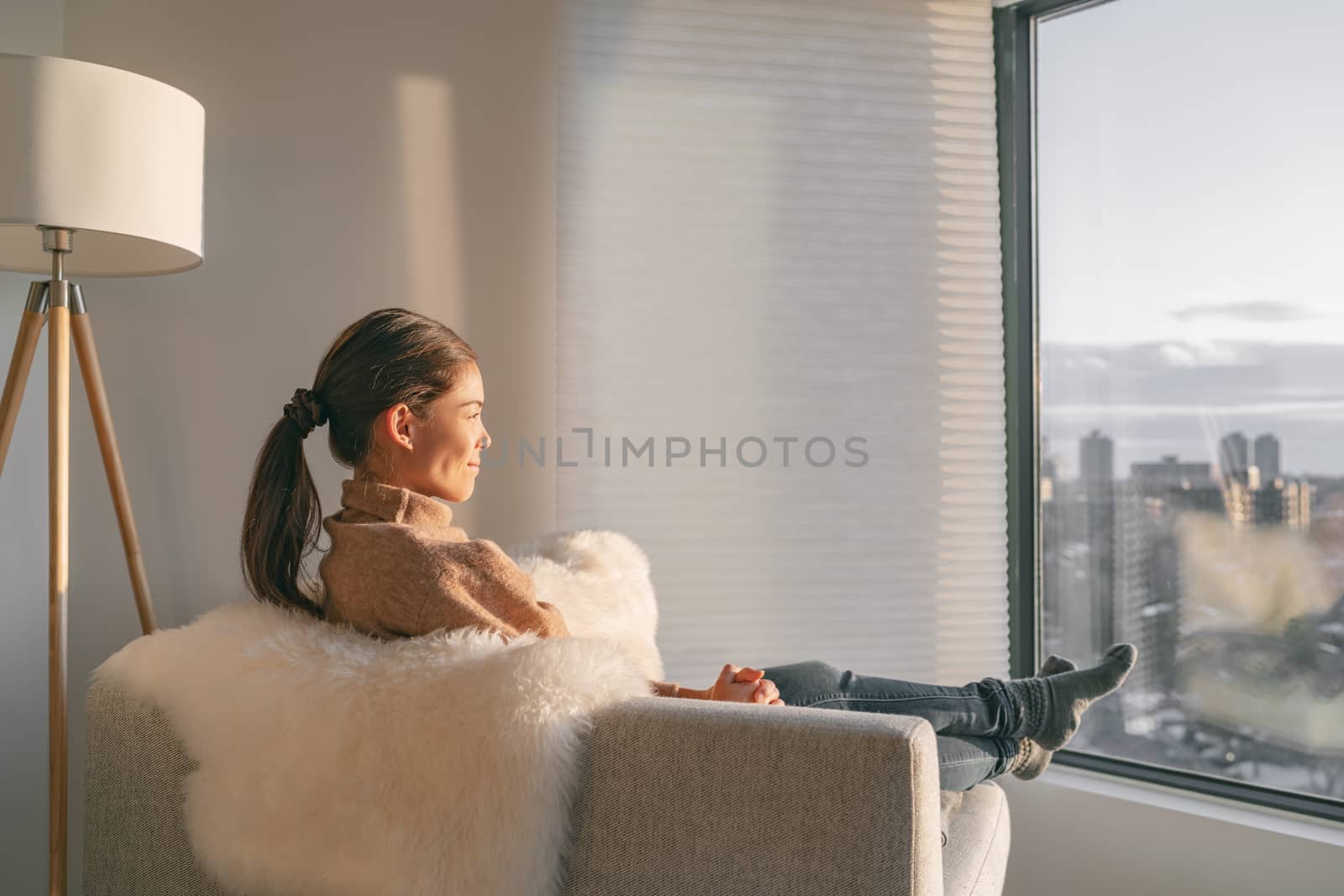 Staying home Asian woman relaxing pensive looking out the window sitting in living room sofa enjoying free time to think and practice mindfulness, indoors during coronavirus outbreak.