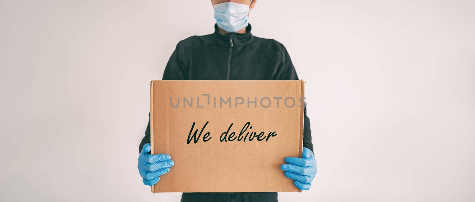 Home delivery WE DELIVER sign on cardboard box banner. Food grocery package online shopping man delivering with gloves and mask for COVID-19 coronavirus social distancing carrying at door.