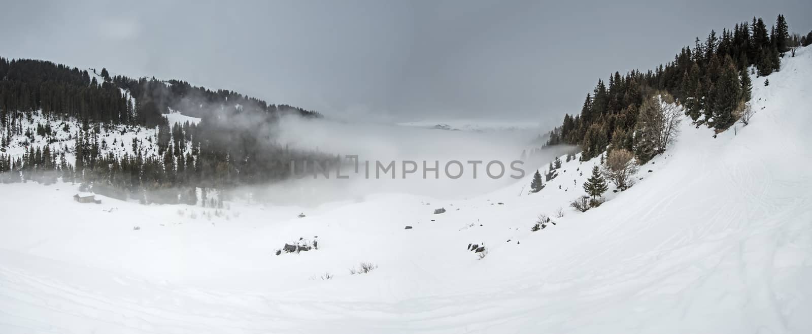 Panoramic view of a snow covered mountain range looking down valley with low clouds and an overcast sky