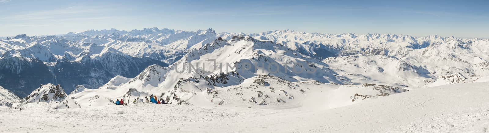 View down a snowy piste with skiers relaxing and view over mountain valley