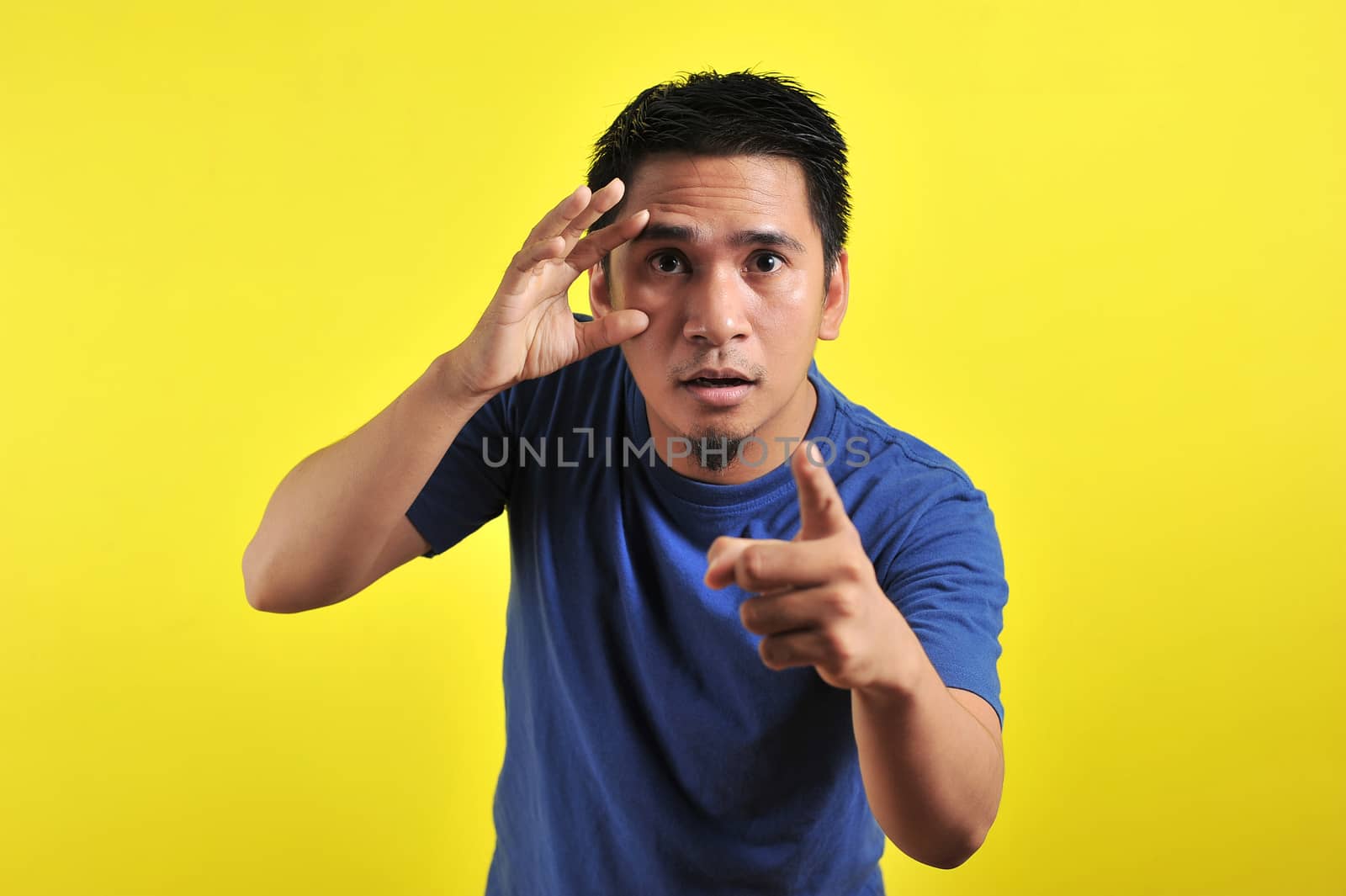 Young Asian man open his eyes and pointing the camera, isolated on yellow background