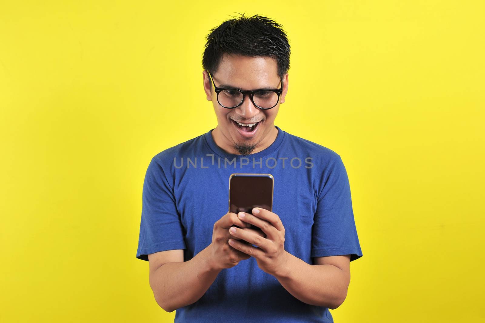 Shocked face of Asian man in blue shirt looking at phone screen on yellow background