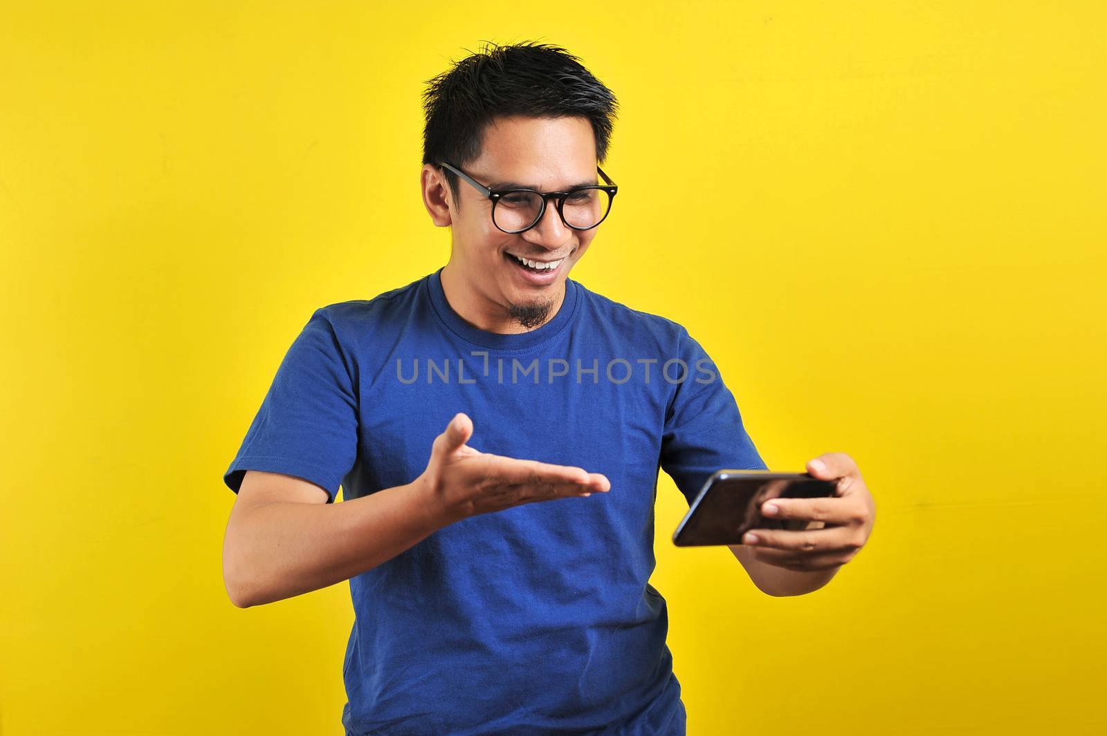 Happy excited Asian man looking at his smartphone and raising his arm up to celebrate success or achievement, isolated on yellow