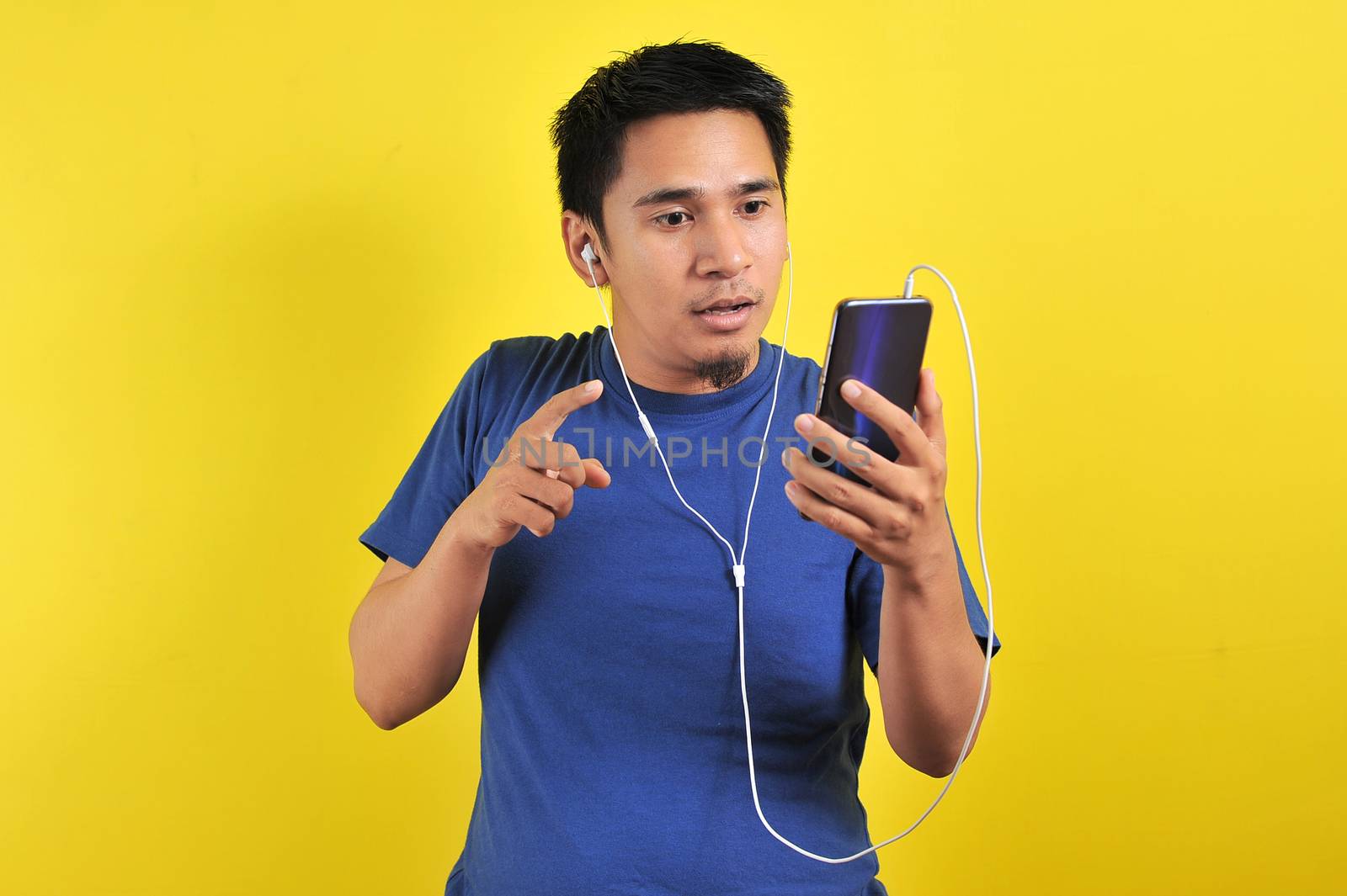Asian man in casual blue t-shirt wearing headset listening to music from smartphone, shock expression, on yellow background