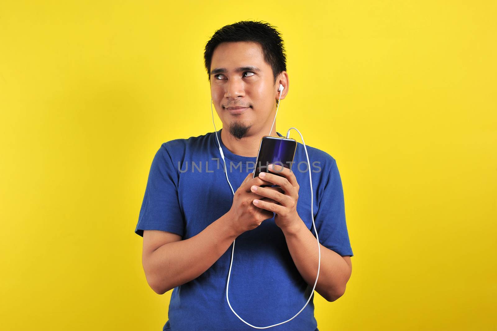 Asian man in casual blue t-shirt wearing headset listening to music from smartphone looking at the copy space, on yellow background.