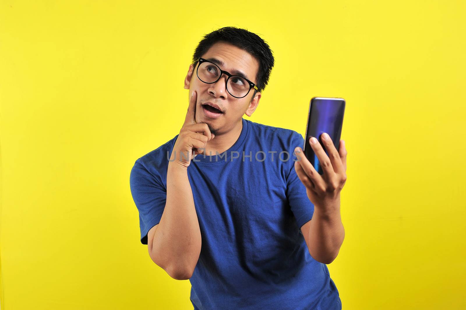 Asian man holding smartphone doing thinking gesture, isolated on yellow background