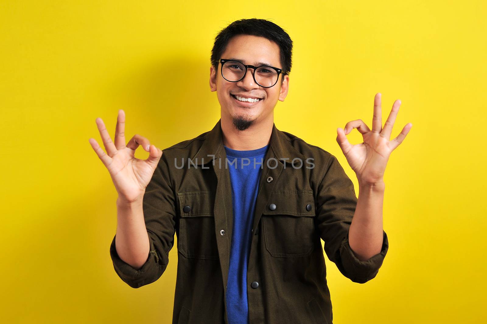 Indonesian man doing double okay wearing eyeglasses, Asian man smiling doing double okay gesture, isolated on yellow
