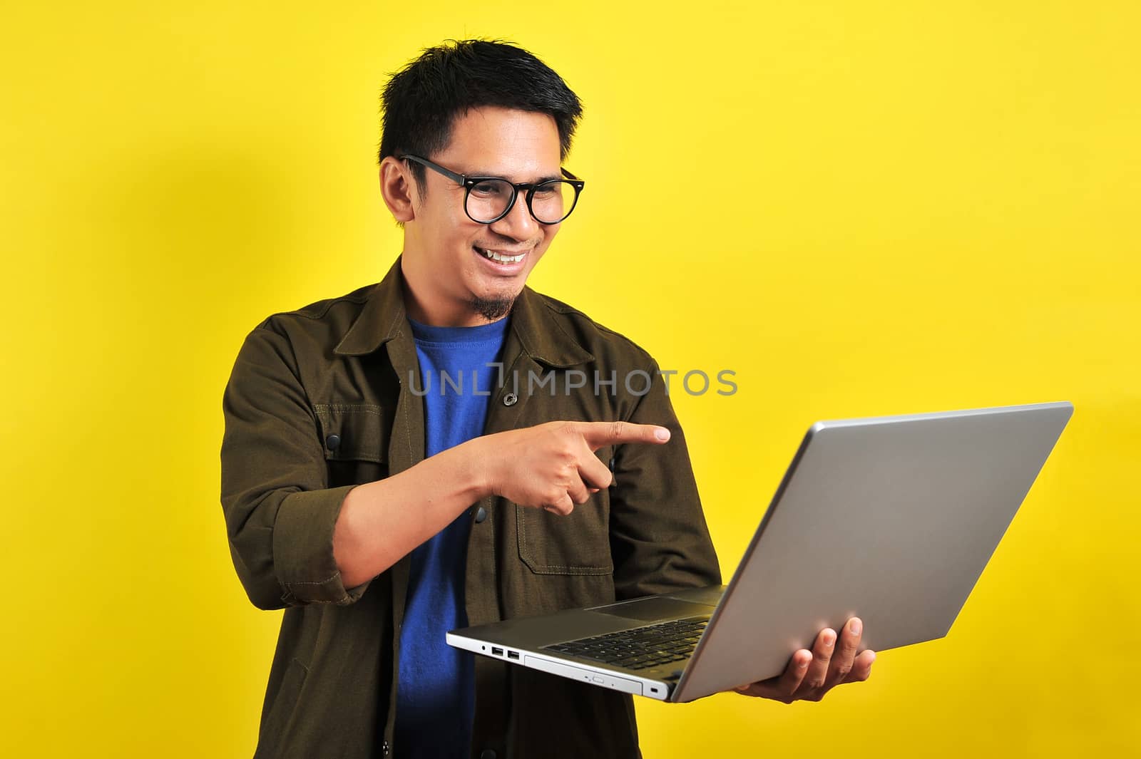 Smiling pleased handsome asian young male entrepreneur, pointing laptop display, isolated on yellow