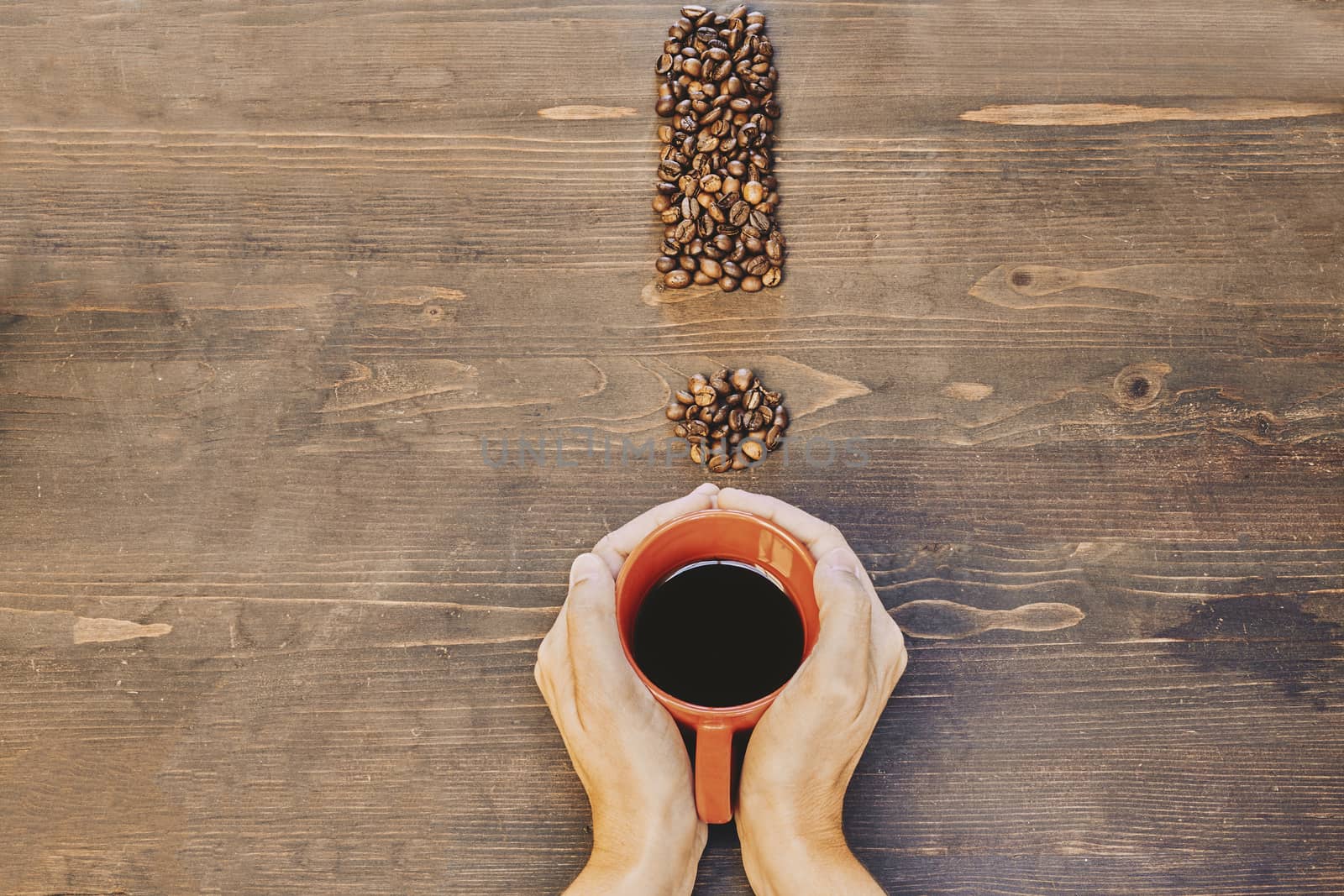 Hands holding a red coffee cup on a wooden table and with an exclamation mark made from coffee beans