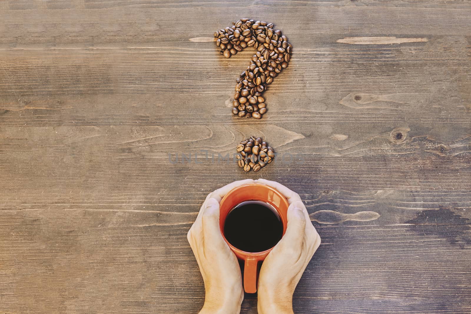 Hands holding a red coffee cup on a wooden table and with a question mark made of coffee beans by Daniel_Mato