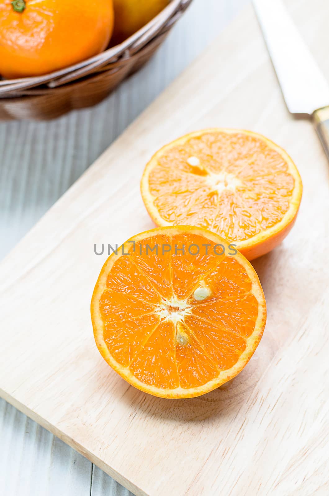 A sliced orange by knife on the wood block and nearby a lot of orange in the wicker brown basket on the white wood table.