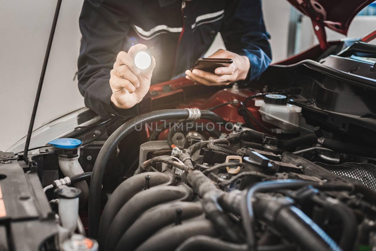 Asian Man mechanic inspection Shine a torch on car engine checking bug in engine from application smartphone. Red car for service maintenance insurance with car engine.