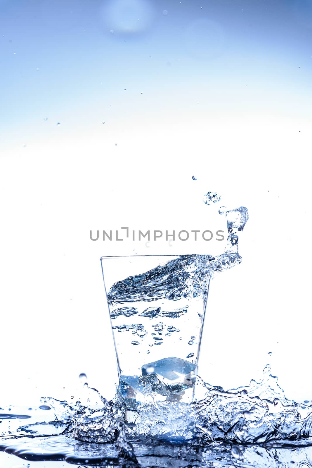 Ice dropping and water splashing in the glass with reflection on the mirror table and isolated on white background.
