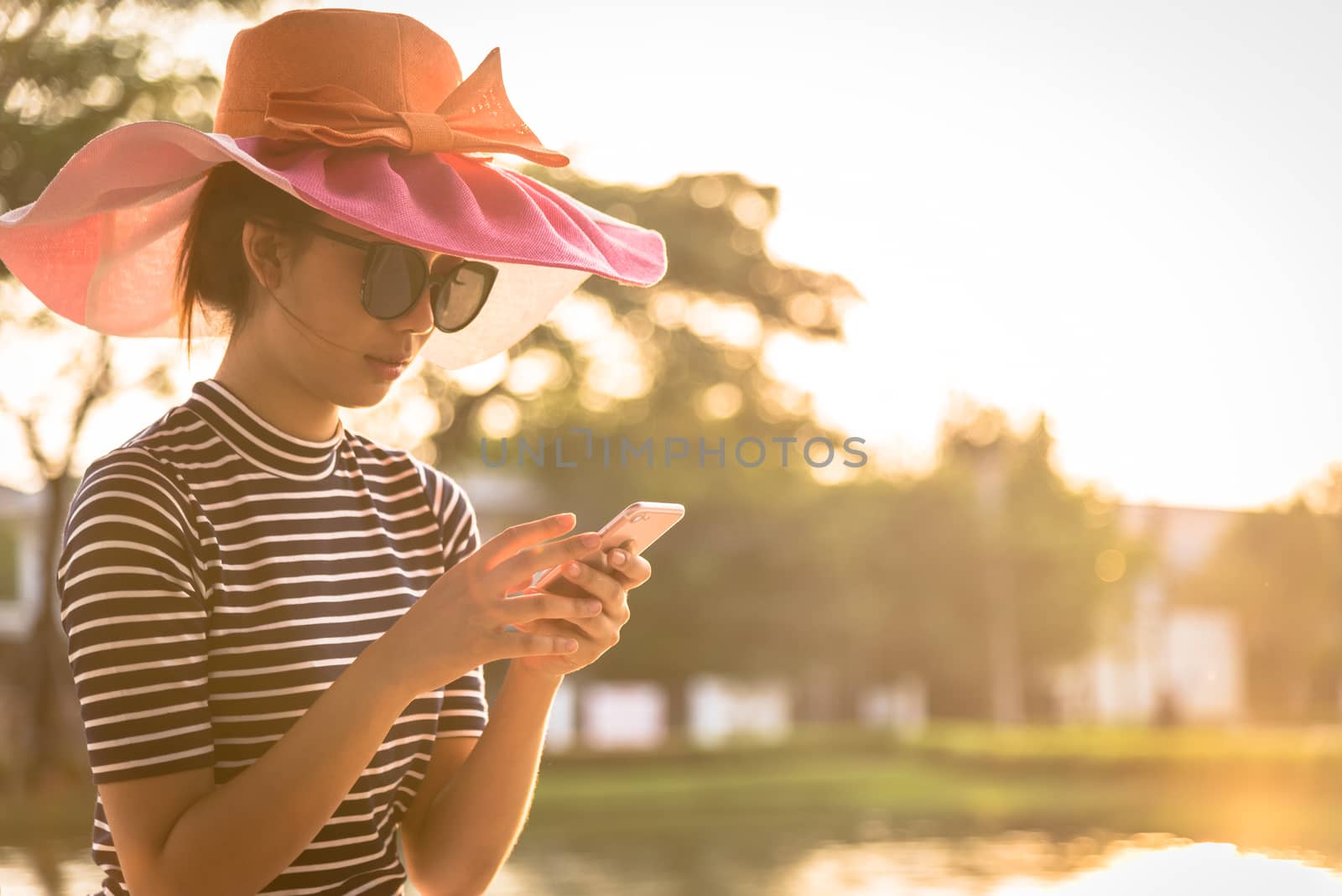 A beautiful woman with pink hat and black eyeglasses has playing phone in the evening with sunset time at garden park and river.