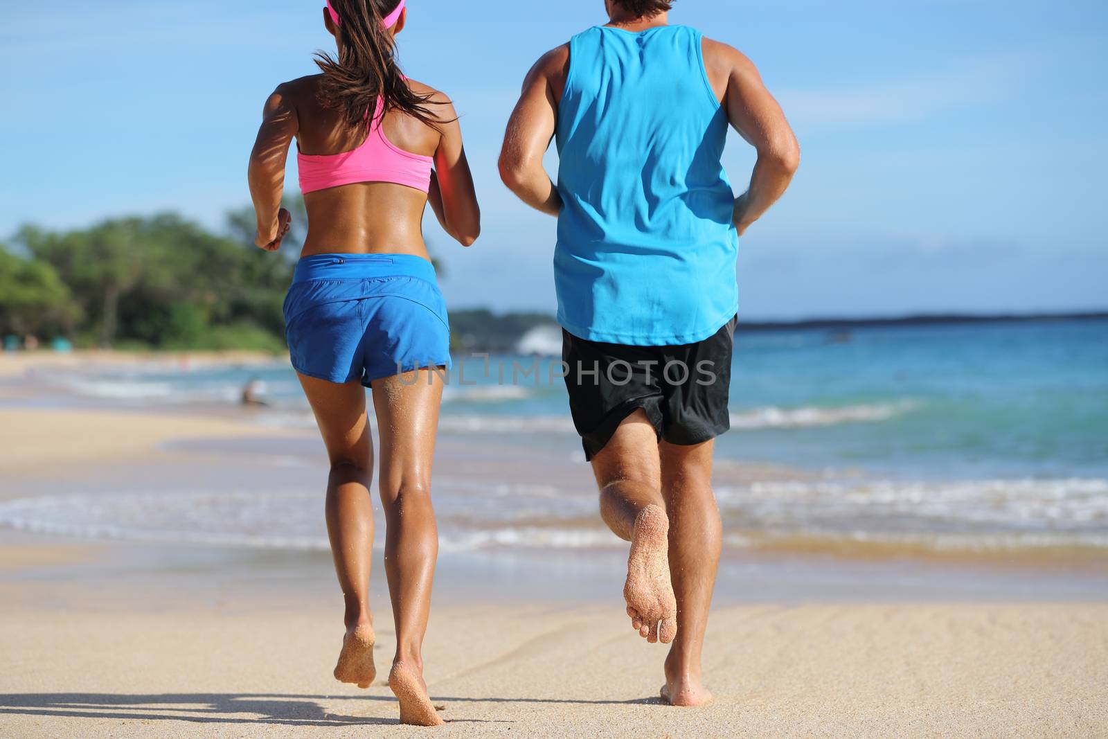 Two athletes runners couple running together on beach. People from behind jogging away barefoot on sand on tropical travel destination. Lower body, legs, feet.