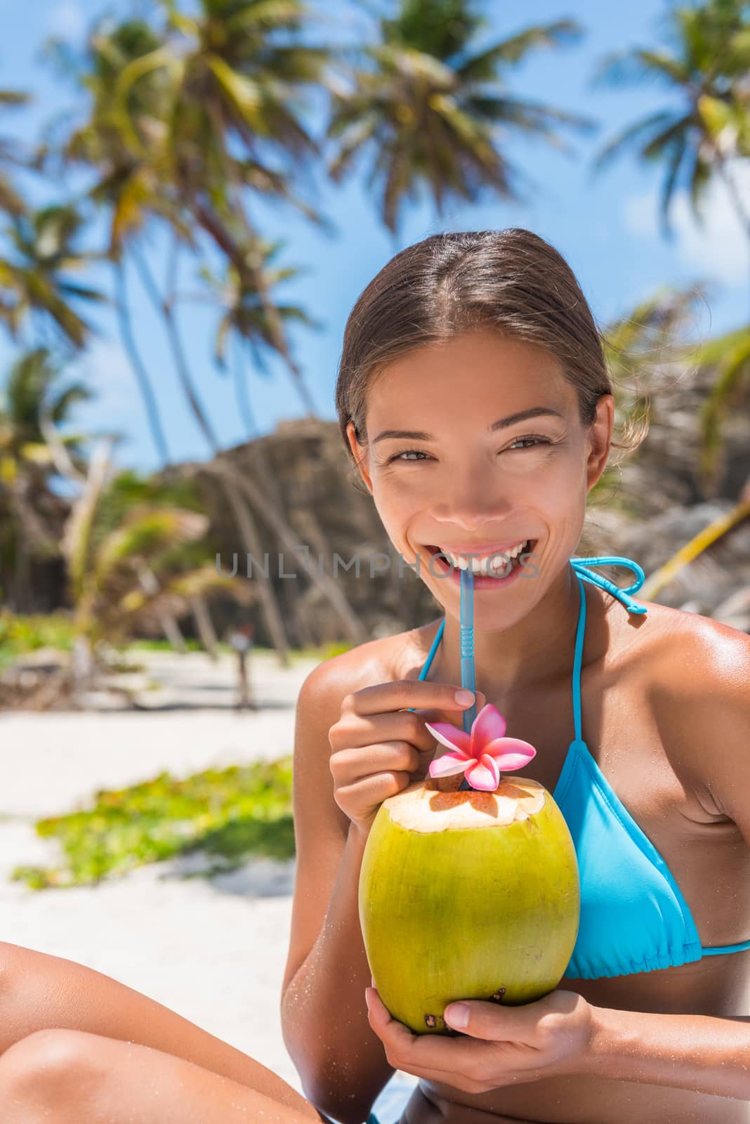 Beautiful Asian bikini woman drinking fresh coconut water directly from fruit with straw on beach. Multiracial girl smiling at camera portrait with perfect teeth sipping on hydration juice food by Maridav