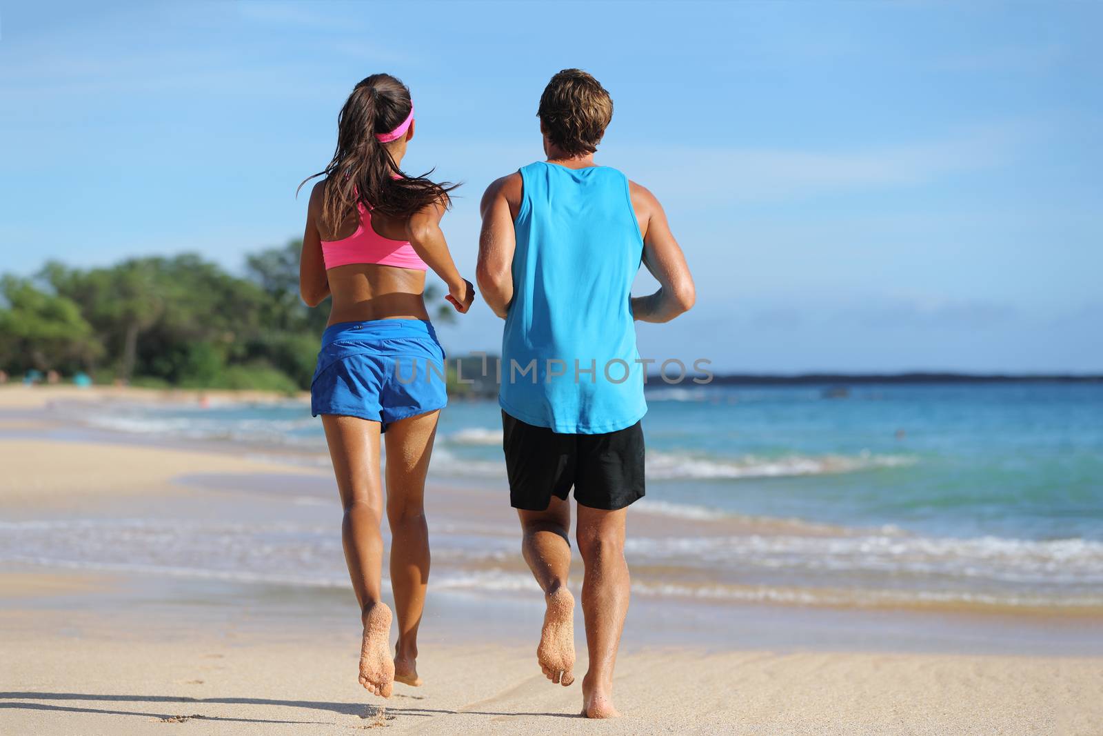 Two fitness athletes running together on beach. People from behind jogging away barefoot on sand on tropical travel destination. Healthy fit young adults with muscular slim legs training cardio.