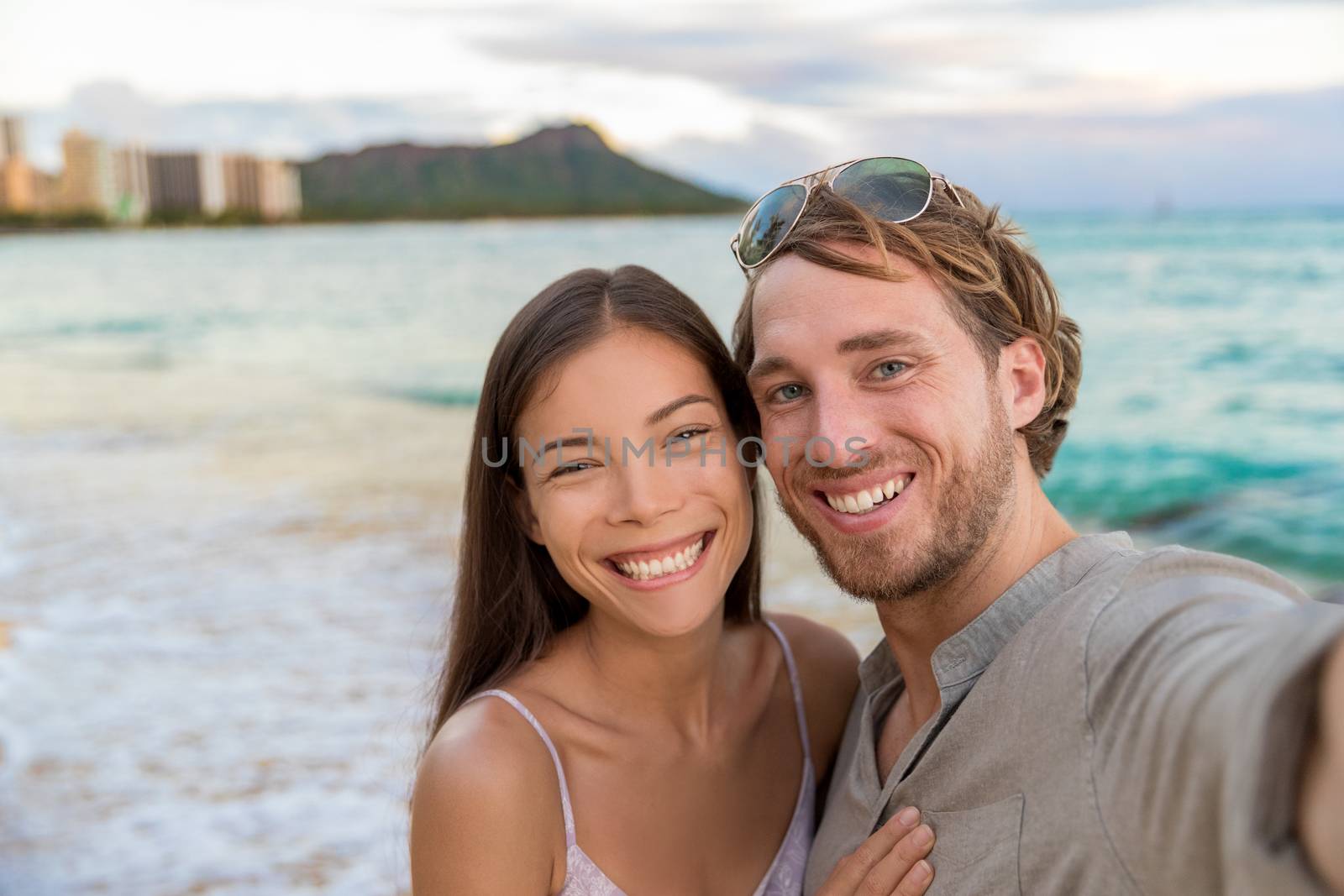 Selfie couple on Waikiki beach taking pictures with smartphone during night out walk on beach summer vacations in Honolulu, Hawaii. Travel destination. Young people having fun on hawaiian holidays by Maridav