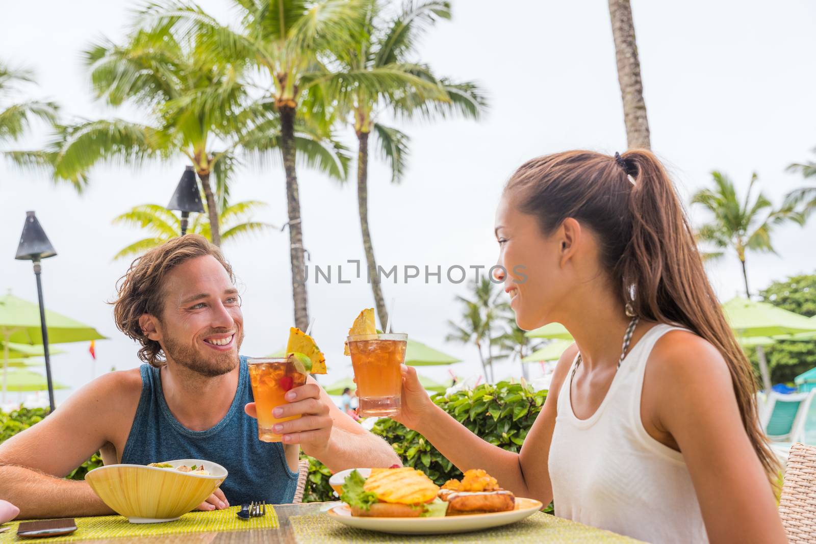 Couple eating at hotel restaurant on Hawaii travel vacation beach drinking hawaiian drink mai tai. Happy people toasting cheers with cocktails. Summer holidays at resort by Maridav